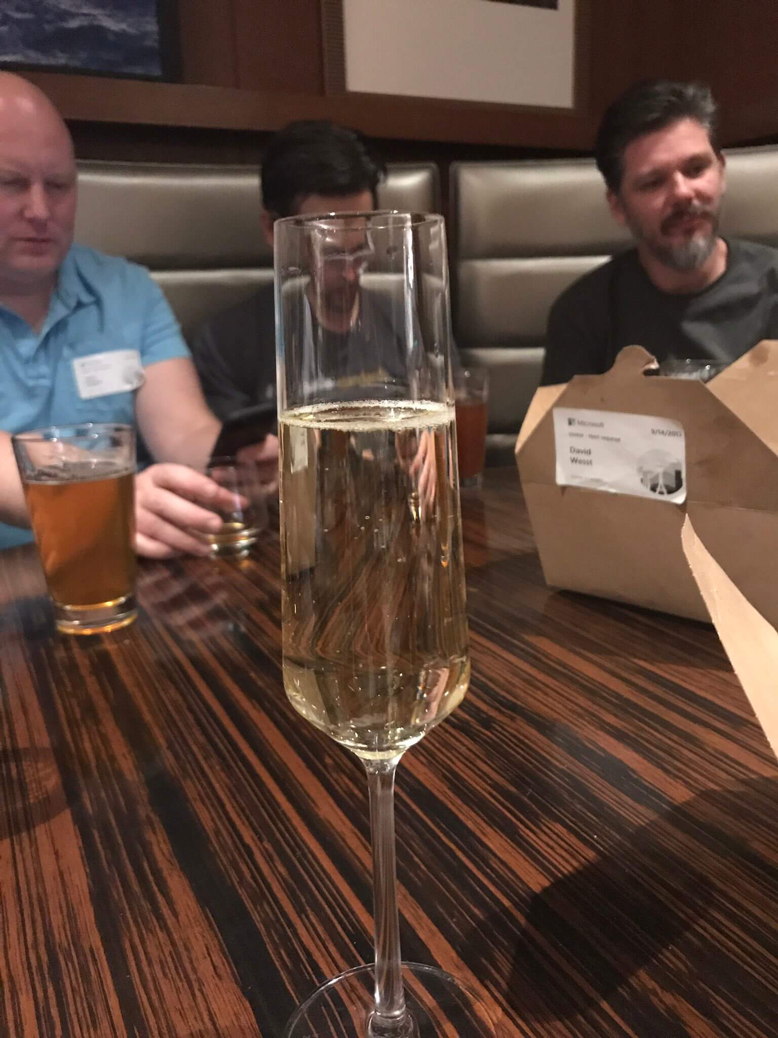 A mostly full Champagne flute in the foreground, a cardboard takeaway container behind it, and blurry people sitting in a booth in the background.