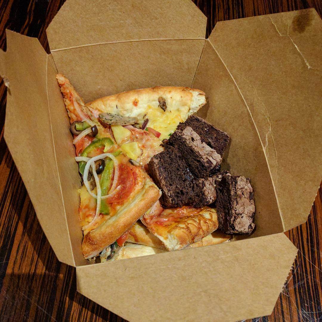 An opened cardboard takeaway container filled to the top with thin slices of pizza and a pile of brownies.