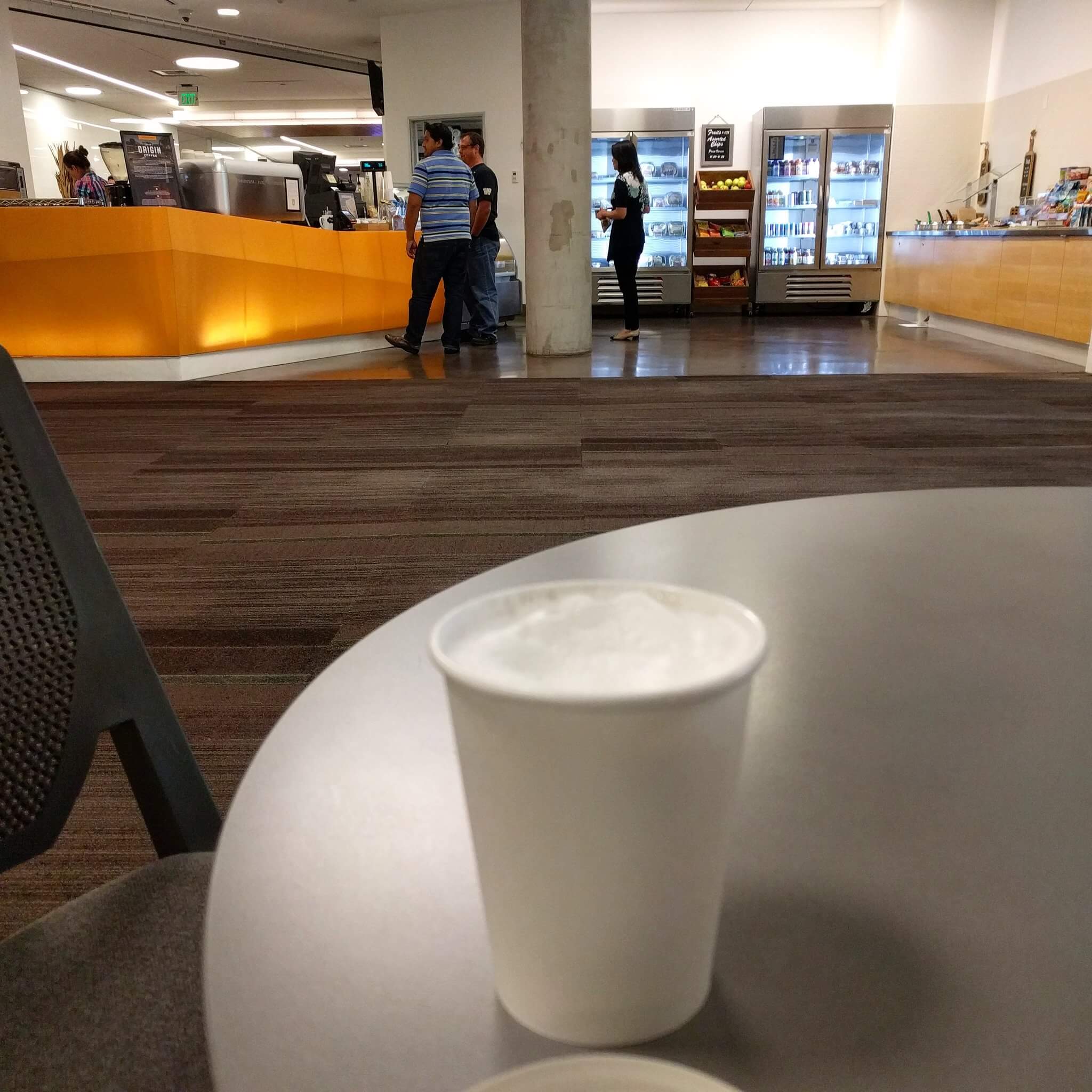 Cappuccino in a paper cup in the foreground, coffee bar and drink cases in the far background, a gulf of carpet between us.