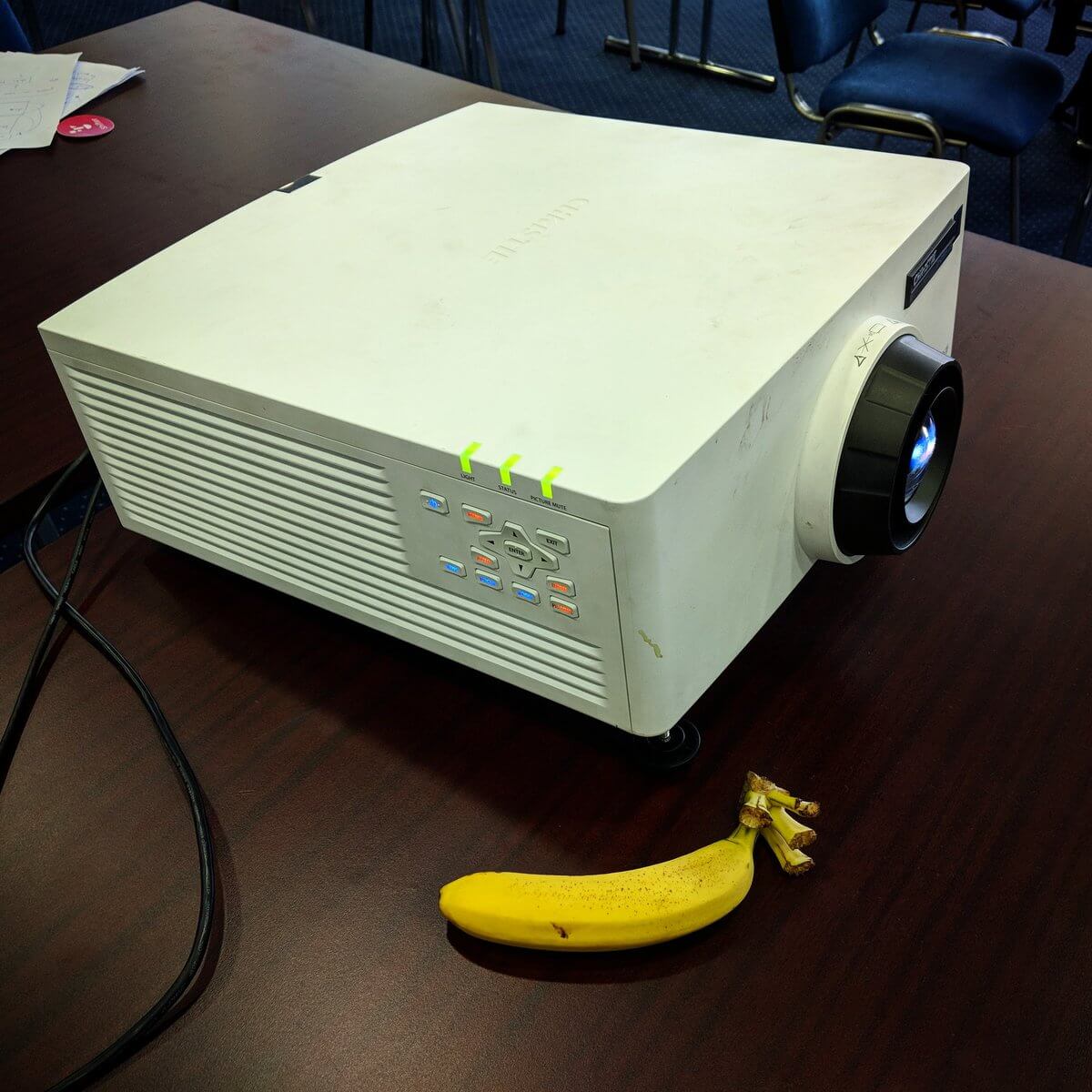 A large computer projector with a worn cover.  It is about a foot-and-a-half square and two-thirds as tall. It has a bank of status lights on one side. Beside it is a banana, dwarfed by the projector.