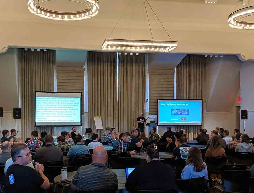 Looking across a room of about 200 people to a stage at the far end when two screens. A slide showing my name and logo, Adrian Roselli LLC, is showing a few feet away from the screen showing the captions where the organizer is thanking me for helping by sponsoring the captions.