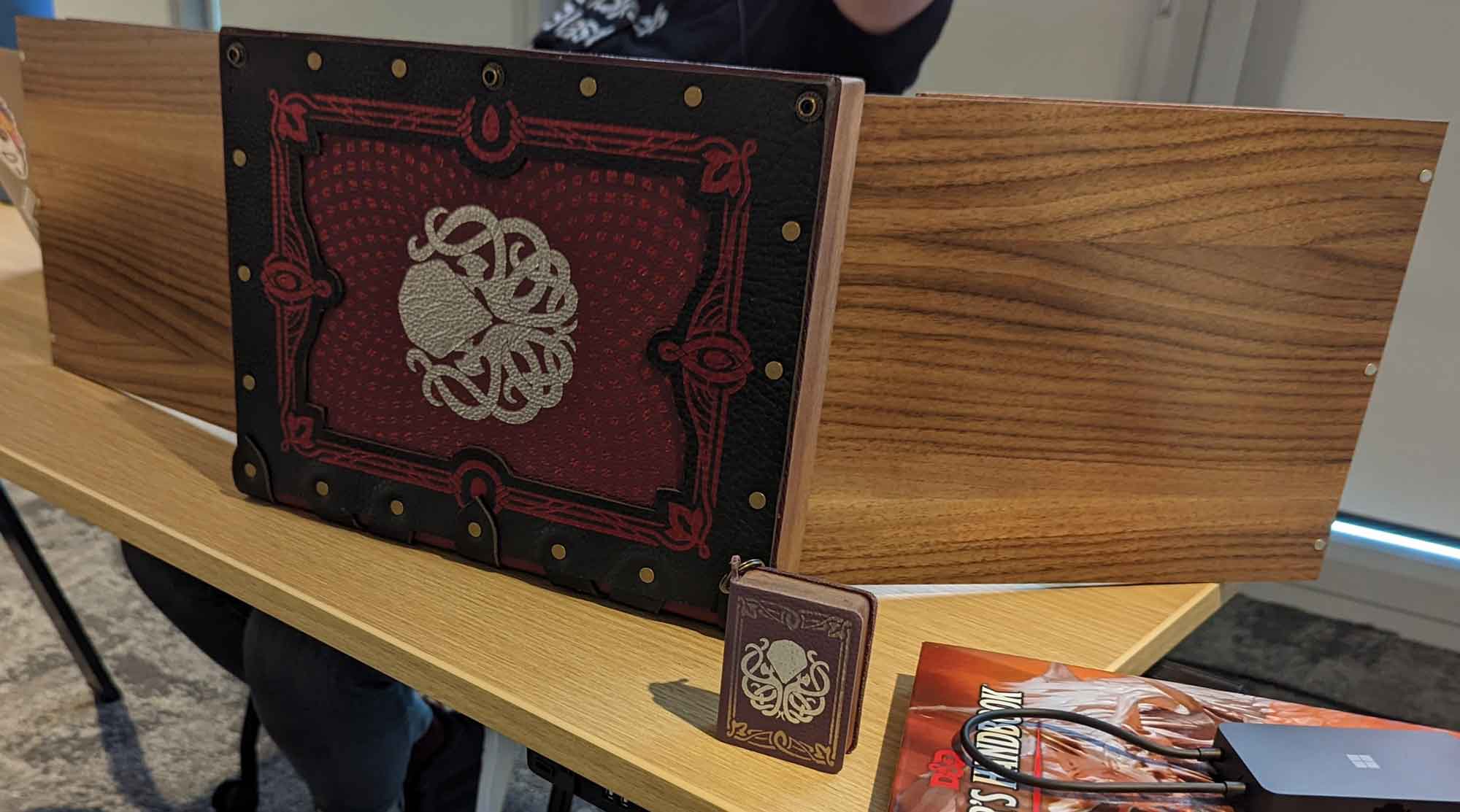 A wooden box large enough to hold D&D books, opened on its side with wooden wings extended; the face of the box is black leather border, read leather center, red foil detail on the edges, and a silver foil cthulhu in the middle.